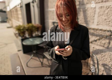 Frau in schwarzem Blazer, die ihr Smartphone hält, auf dem Bürgersteig der Stadt steht. Moderne Mode, urbaner Lifestyle, Nahaufnahme, lässige Eleganz. Stockfoto