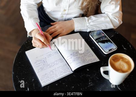 Junge Frau, die in einem Notizbuch an einem gemütlichen Café-Tisch mit Kaffee und Smartphone schreibt. Konzentriert und nachdenklich plant oder studiert sie in einer ruhigen, stilvollen Umgebung Stockfoto