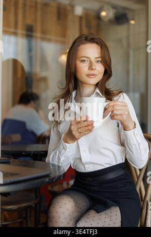 Schöne Frau in weißem Hemd, die Kaffeepause in stilvollem Café genießt. Gemütliche Atmosphäre, Tageslicht und modernes Lifestyle-Konzept. Stockfoto