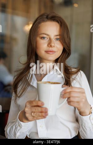 Selbstbewusste Geschäftsfrau, die in einem Café Kaffee trinkt. Eine ungezwungene Arbeitsumgebung, sanftes Licht und freundlicher Ausdruck spiegeln einen entspannten, professionellen Lebensstil wider Stockfoto