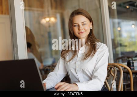 Selbstbewusste junge Geschäftsfrau in einem weißen Hemd, die in einem modernen Café an einem Laptop arbeitet. Professionell und stilvoll, sie lächelt, während sie an einem hölzernen Ta sitzt Stockfoto