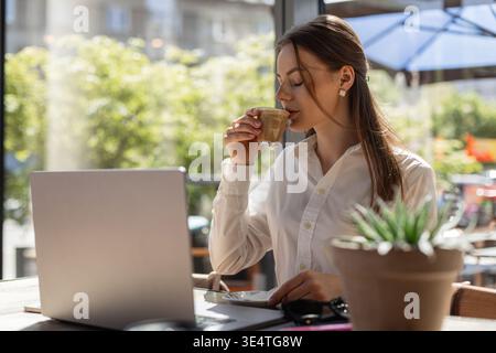 Selbstbewusste Frau genießt Kaffee und arbeitet an einem Laptop in einem sonnigen Café. Freiberuflicher und externer Arbeitsstil. Stockfoto