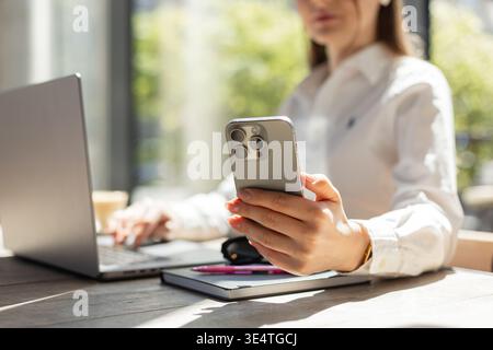 Geschäftsfrau, die an einem Laptop arbeitet und das Smartphone in der Hand hält. Helles, modernes Büro mit Tageslicht. Konzept für Fernarbeit, Kommunikation und Profi Stockfoto