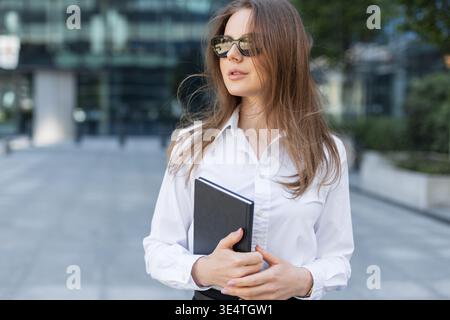 Stilvolle junge Frau in formellem Hemd und Sonnenbrille mit Notizbuch in einem Geschäftsviertel der Stadt. Selbstbewusster und professioneller Look. Stockfoto