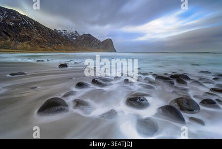 Wasser fließt über riesige Felsbrocken im Sand am Unstad Strand, Unstad. Langbelichtungsbild. Atemberaubender Sonnenuntergang mit Bergen am Horizont auf Lofote Stockfoto