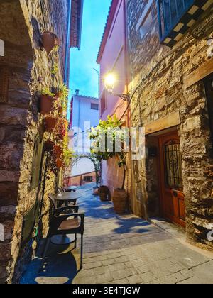 Enge kopfsteingepflasterte Gasse in Sueras, castellon, schlängelt sich zwischen Steinhäusern mit leuchtenden Straßenlampen, Topfpflanzen und Cafés im Freien Stockfoto