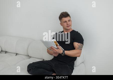 Stilvoller, hübscher Geschäftsmann in schwarzer Kleidung, der ein Magazin liest und auf dem weißen Sofa im Zimmer relaxt. Coole Guy Rest Stockfoto