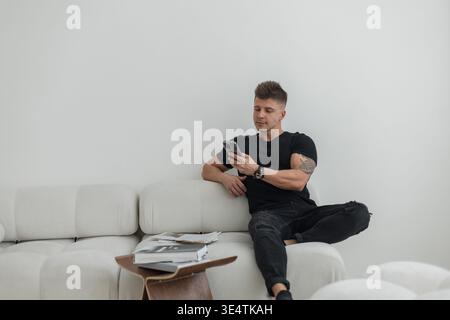 Cooler erfolgreicher gutaussehender Mann in schwarzer Kleidung, der auf dem Sofa sitzt und ein Smartphone hält, das Nachrichten online liest Stockfoto