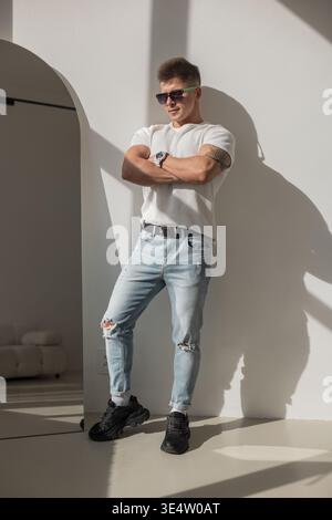 Selbstbewusster junger Mann in lässigem Outfit und Sonnenbrille posiert im hellen Sonnenlicht. Mit weißem Hemd, zerrissener Jeans, schwarzen Turnschuhen und einer Uhr, stehend Stockfoto