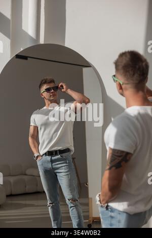 Tätowierter Mann in weißem T-Shirt und Jeans, der sein Spiegelbild im Spiegel ansieht. Helles, modernes Interieur, Sonnenlicht wirft Schatten. Selbstbewusst und stilvoll Stockfoto