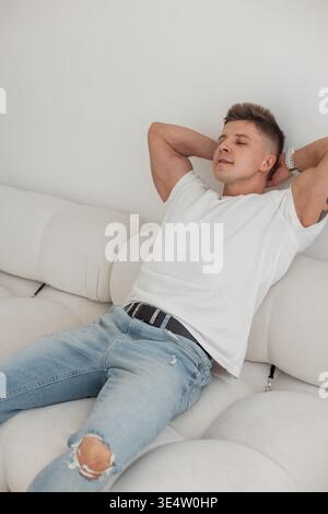 Stilvoller Mann in lässigem Outfit, der sich auf einem weißen Sofa entspannt, Arme hinter dem Kopf. Moderne Inneneinrichtung, gemütliche und selbstbewusste Atmosphäre. Stockfoto