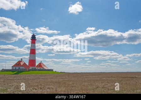 Ein hoher rot-weiß gestreifter Leuchtturm steht neben Häusern auf einem grünen grasbewachsenen Feld mit einer trockenen Wiese und einem blauen Himmel voller verstreuter Wolken. Stockfoto