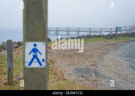 Eine nebelige Szene am See mit einem hölzernen Pier, Schotterpfad, Holzpfosten und einem blauen Fußgängerschild an einem nebeligen Tag am Wasser. Stockfoto