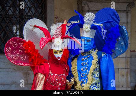 Ein paar venezianische Masken mit aufwendigen Kostümen in Rot und Königsblau reich an Federn, Blumen und goldenen Details historischer architektonischer Hintergrund Stockfoto