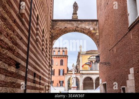 Perspektivischer Blick durch den Arco della Costa in Richtung Piazza dei Signori in Verona. Im Vordergrund befinden sich Mauern aus Ziegelstein und Tuffstein, während Dantes Statue und Stockfoto