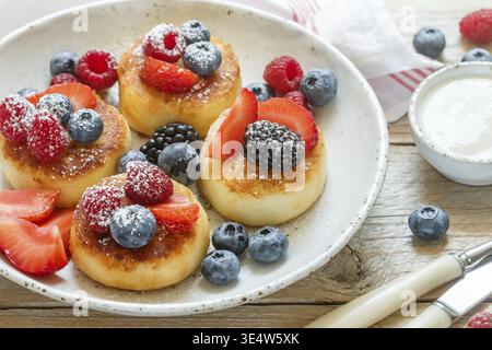 Essen, Hüttenkäsepfannkuchen, Syrniki, Quark-Krapfen mit frischen Beeren (Himbeere, Erdbeere, Heidelbeere, Brombeere) und Puderzucker in einem weiß Stockfoto