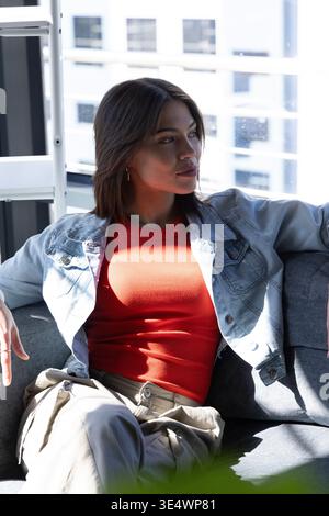 Frau, die auf einer grauen Couch in der Nähe eines großen Fensters sitzt, eine Jeansjacke und ein rotes Oberteil trägt Stockfoto