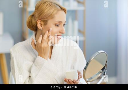 Lächelnde junge Frau im Bademantel, die Gesichtscreme aufträgt und in den Spiegel blickt während der Hautpflegeroutine. Stockfoto