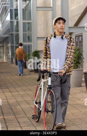 Asiatischer Mann schiebt weißes Rennrad mit roten Felgen, während er Mütze und Kopfhörer auf dem Flur trägt Stockfoto