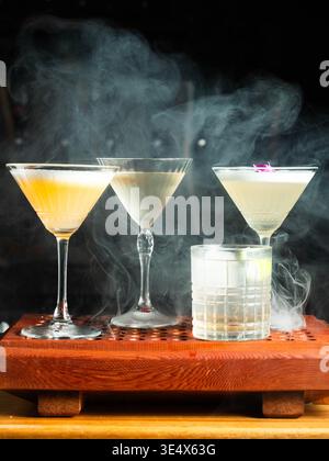 Drei Cocktails dämpfen sanft auf einem Holztablett. Jedes Getränk variiert in Farbe und Stil, was Intrigen schafft. Rauchen Sie Locken um die Brille, um die zu verstärken Stockfoto