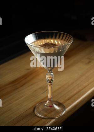 Silberner Cocktail glänzt in Martini-Glas. Die Holzoberfläche sorgt für einen warmen, rustikalen Kontrast. Glas hält Flüssigkeit mit dezent schimmerndem Licht. Dunkler Hintergrund e Stockfoto