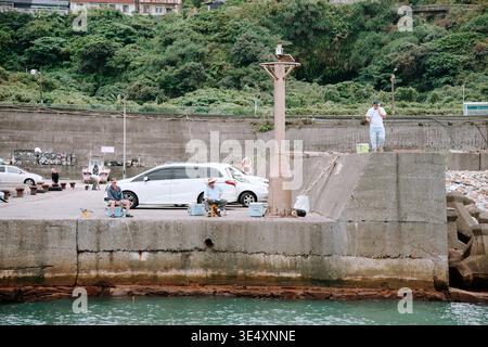 New Taipei City, Taiwan - 30. Oktober 2023: Drei Männer fischen an einem warmen Tag entlang eines Betonhafens, mit grünen Hügeln und geparkten Autos dahinter. Stockfoto