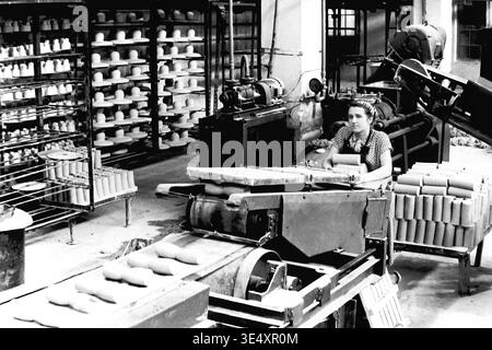 Sloviansk, 1950er-1960er Jahre, Armature-Insulator Plant (AIZ). Eine Fabrikarbeiterin betreibt ein mechanisches Fördersystem, das rohe zylindrische Keramikrohlinge verarbeitet. Das Schwarzweiß-Archivfoto erfasst die aktive industrielle Produktionsumgebung mit umfangreichen Trockenregalen, gefüllt mit geformten Hochspannungs-Isolatoren, die im Hintergrund sichtbar sind. Dieses authentische Bild unterstreicht die entscheidende Rolle der Frauen in der sowjetischen Massenproduktion und bewahrt das technische Erbe und das Arbeitsleben des friedlichen Donbas. Stockfoto