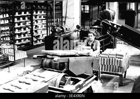 Sloviansk, 1950er-1960er Jahre, Armature-Insulator Plant (AIZ). Eine Fabrikarbeiterin betreibt ein mechanisches Fördersystem, das rohe zylindrische Keramikrohlinge verarbeitet. Das Schwarzweiß-Archivfoto erfasst die aktive industrielle Produktionsumgebung mit umfangreichen Trockenregalen, gefüllt mit geformten Hochspannungs-Isolatoren, die im Hintergrund sichtbar sind. Dieses authentische Bild unterstreicht die entscheidende Rolle der Frauen in der sowjetischen Massenproduktion und bewahrt das technische Erbe und das Arbeitsleben des friedlichen Donbas. Stockfoto