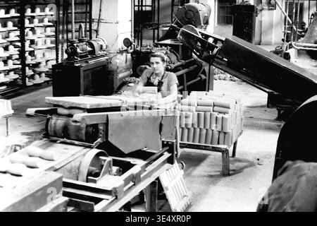 Sloviansk, 1950er-1960er Jahre, Armature-Insulator Plant (AIZ). Eine Fabrikarbeiterin betreibt ein mechanisches Fördersystem, das rohe zylindrische Keramikrohlinge verarbeitet. Das Schwarzweiß-Archivfoto erfasst die aktive industrielle Produktionsumgebung mit umfangreichen Trockenregalen, gefüllt mit geformten Hochspannungs-Isolatoren, die im Hintergrund sichtbar sind. Dieses authentische Bild unterstreicht die entscheidende Rolle der Frauen in der sowjetischen Massenproduktion und bewahrt das technische Erbe und das Arbeitsleben des friedlichen Donbas. Stockfoto