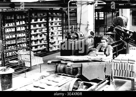 Sloviansk, 1950er-1960er Jahre, Armature-Insulator Plant (AIZ). Eine Fabrikarbeiterin betreibt ein mechanisches Fördersystem, das rohe zylindrische Keramikrohlinge verarbeitet. Das Schwarzweiß-Archivfoto erfasst die aktive industrielle Produktionsumgebung mit umfangreichen Trockenregalen, gefüllt mit geformten Hochspannungs-Isolatoren, die im Hintergrund sichtbar sind. Dieses authentische Bild unterstreicht die entscheidende Rolle der Frauen in der sowjetischen Massenproduktion und bewahrt das technische Erbe und das Arbeitsleben des friedlichen Donbas. Stockfoto