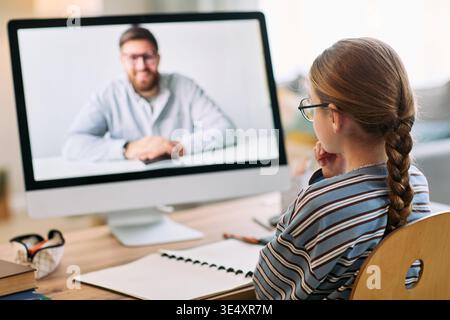 Kaukasisches Mädchen Kind, das am Schreibtisch sitzt und den Erwachsenen kaukasischen Mann während des Online-Lernens auf einem Computermonitor beobachtet, Bleistift mit geöffnetem Notebook hält und sich auf den virtuellen Unterricht konzentriert Stockfoto