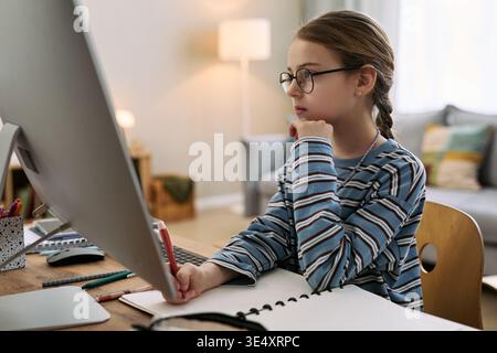 Kaukasisches Mädchen mit Brille am Schreibtisch sitzend am Desktop-Computer, während es im Notebook schreibt, sich auf den Bildschirm konzentriert, studiert oder Hausaufgaben in der häuslichen Umgebung macht Stockfoto