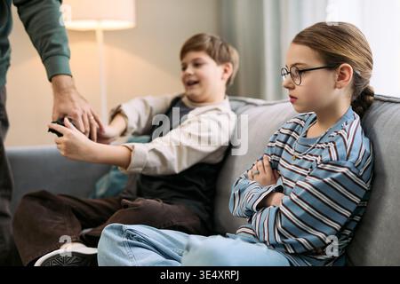 Kaukasischer Junge, der auf dem Sofa sitzt und die Fernbedienung hält, während die erwachsene Hand greift, um sie zu nehmen, kaukasisches Mädchen mit Brille neben ihm sitzt, die Arme kreuzt und unzufrieden wegblickt Stockfoto