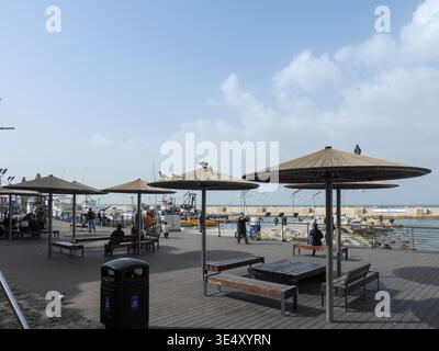 Die Leute laufen und entspannen entlang der Jaffa Port Promenade, sitzen in Cafés im Freien und genießen den Blick auf den Hafen mit Booten, die im Yachthafen angedockt sind Stockfoto