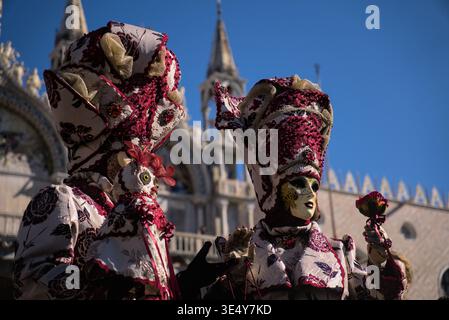 Zwei verzierte venezianische Karnevalsmasken, die mit komplizierten Mustern und Federn verziert sind, stehen vor der berühmten Basilika di San Marco unter einem klaren Blau Stockfoto