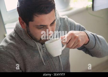 Mann in grauem Kapuzenpullover, der heissen Kaffee aus einer weißen Tasse in der Nähe des Fensters trinkt Stockfoto