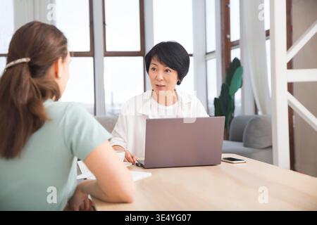Glückliche, multiethnische Kollegen sitzen zusammen am Coworking-Platz, erfahrenere Mitarbeiter teilen sich. Die entzückende asiatische Geschäftsfrau berät den Kunden im Büro || Model veröffentlicht Stockfoto