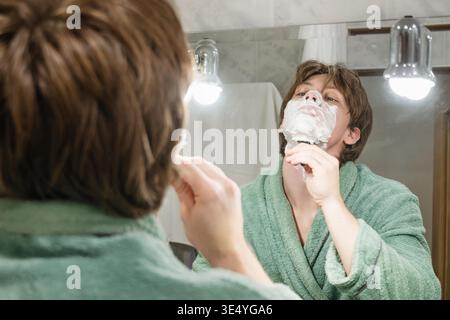 Junger Mann rasiert sich das Kinn mit grünem Bademantel im Badezimmerspiegel. Rasierroutine am Morgen || Modell veröffentlicht Stockfoto