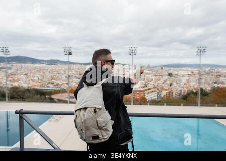 Schöner Reisender Mann mit Sonnenbrille in schwarzer Jacke mit Rucksack auf Aussichtsplattform mit Schwimmbad und Zeigefinger auf Barcelona c Stockfoto