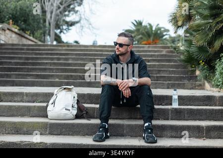 Stilvoller, schöner Reisender mit Sonnenbrille und Hoodie mit Sneakers auf Stufen mit Rucksack im Park in Barcelona Stockfoto