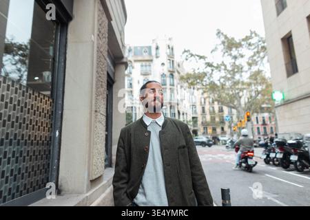 Glücklicher, gutaussehender Mode-Urban-Mann in trendigen modischen Kleidern mit Mantel, der in Barcelona spaziert Stockfoto