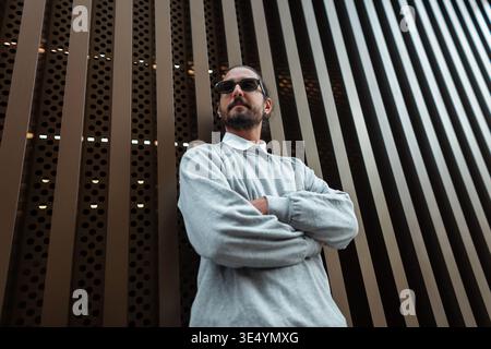 Stilvoller, schöner Modemann mit modischer Sonnenbrille in stilvollem Sweatshirt, der neben Bronze-Metallwand auf der Straße steht Stockfoto