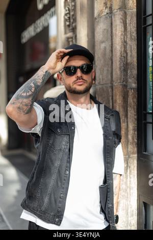 Ein cooler, gutaussehender Mann in modischer Kleidung mit Sonnenbrille trägt eine schwarze Kappe und spaziert durch Barcelona Stockfoto