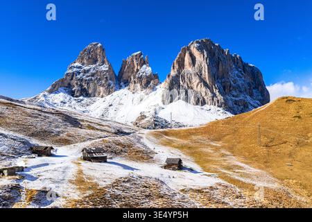 Aus der Vogelperspektive durchdringen schneebedeckte Gipfel den azurblauen Himmel und werfen Schatten auf die rustikalen Holzhütten auf den goldenen, frostigen Hängen, dem Langkofel Sella Stockfoto