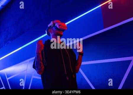 Cooler trendiger Mann in vr-Brille, der 360°-Videos mit Virtual-Reality-Headset auf dunklem Hintergrund mit Neonlicht ansieht. Der Kerl hält einen Joystick und pla Stockfoto