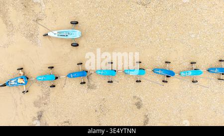 Aus der Vogelperspektive auf eine Reihe von Strandyachten am Strand, deren leuchtend blaue Körper im Kontrast zum braunen Sand stehen, Fort-Mahon-Plage, Hauts-de-France Stockfoto