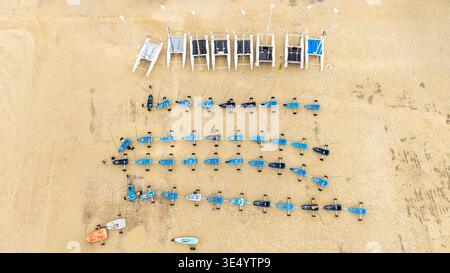 Aus der Vogelperspektive sehen Sie Katamarane und Windsurfbretter am Sandstrand, einen geometrischen Wandteppich aus Blau und weiß vor der goldenen Küste Fort-Ma Stockfoto