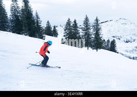 Blick auf einen Skifahrer in leuchtend orangefarbener Jacke, der eine unberührte, schneebedeckte Piste unter aufragenden Evergreens hinabschneidet, Val-d Illiez, Wallis, Schweiz. Stockfoto