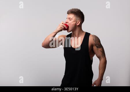 Cooler, gutaussehender sportlicher starker Mann mit muskulösem Körper mit schwarzem Tanktop, der Apfel auf grauem Hintergrund isst. Gesunde Lebensmittel und Vitamine Stockfoto