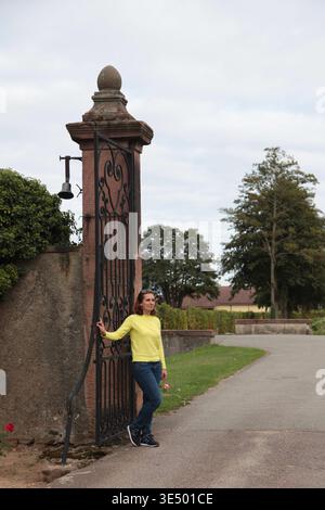 Frau mit gelbem Pullover und Jeans lehnt sich an ein kunstvoll verziertes Eisentor in einem Garten, mit einem Weg und Bäumen im Hintergrund Stockfoto